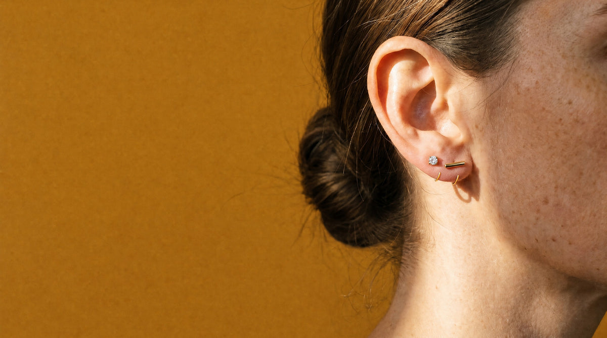 Clear Round Open Hoop and Bar Open Hoop in 18K Gold Vermeil on an ear in profile, against mustard paper in a beam of sun.