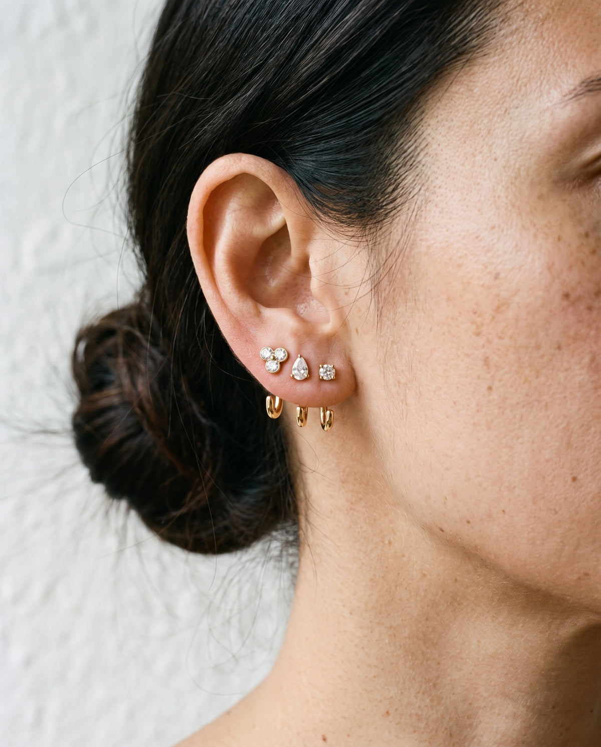 The Clear Cluster, Teardrop and Round set in 18K Gold Vermeil on one ear, in window light against a bone wall.