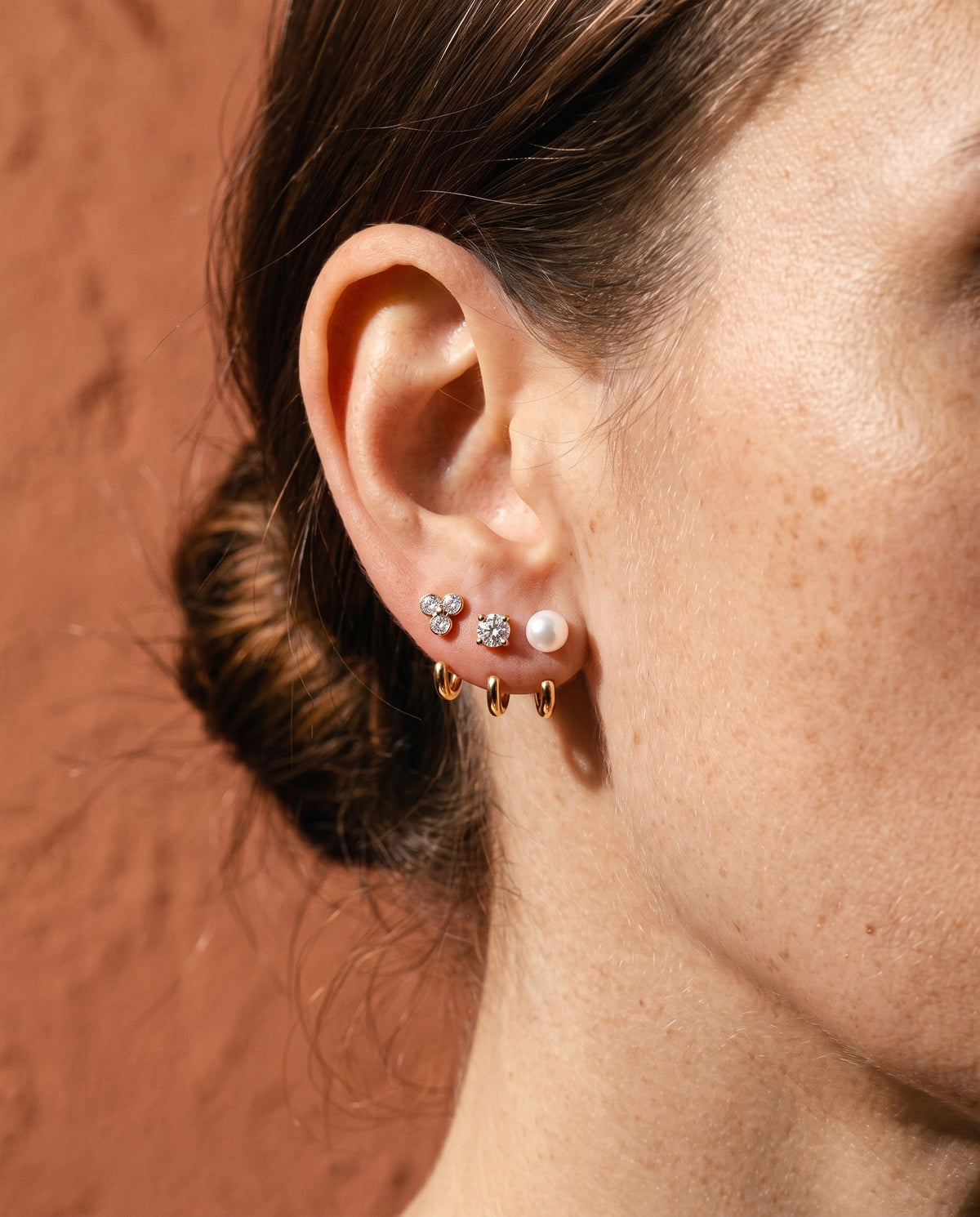 The Clear Cluster, Round and White Bead set in 18K Gold Vermeil on one ear, against a terracotta wall.