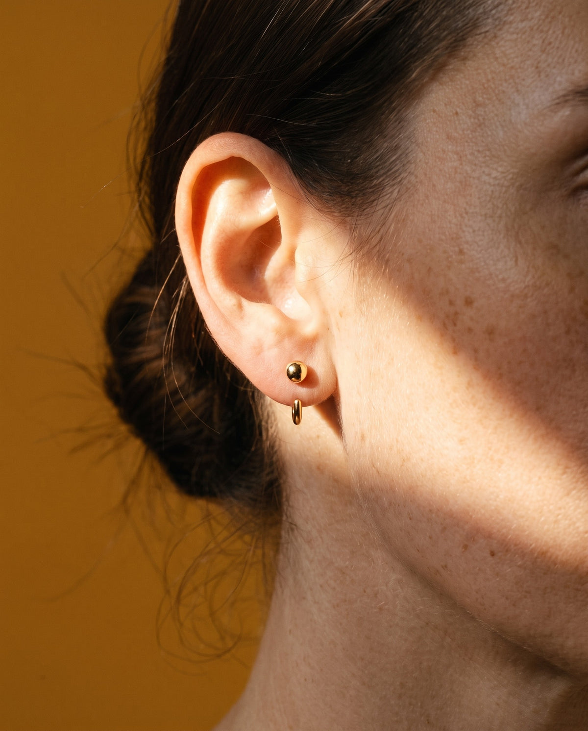 Ball Open Hoop in 18K Gold Vermeil on an earlobe, in a beam of sun on mustard paper.
