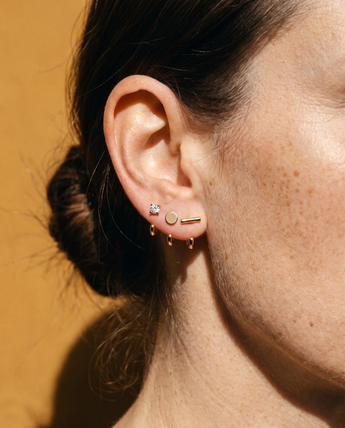 Clear Round, Disc and Bar Open Hoops in 18K Gold Vermeil on one ear, against mustard paper.