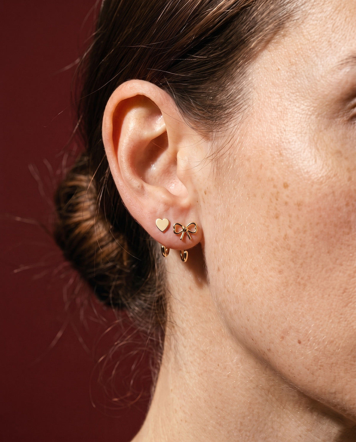 Heart Open Hoop and Bow Open Hoop in 18K Gold Vermeil on one ear, against dark red paper.